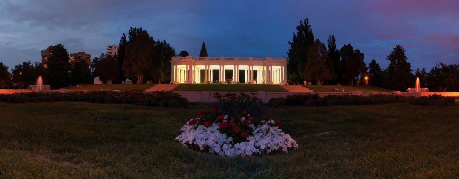 The marble pavilion at Cheesman Park lit up against a deep blue dusk sky, with flower beds in front