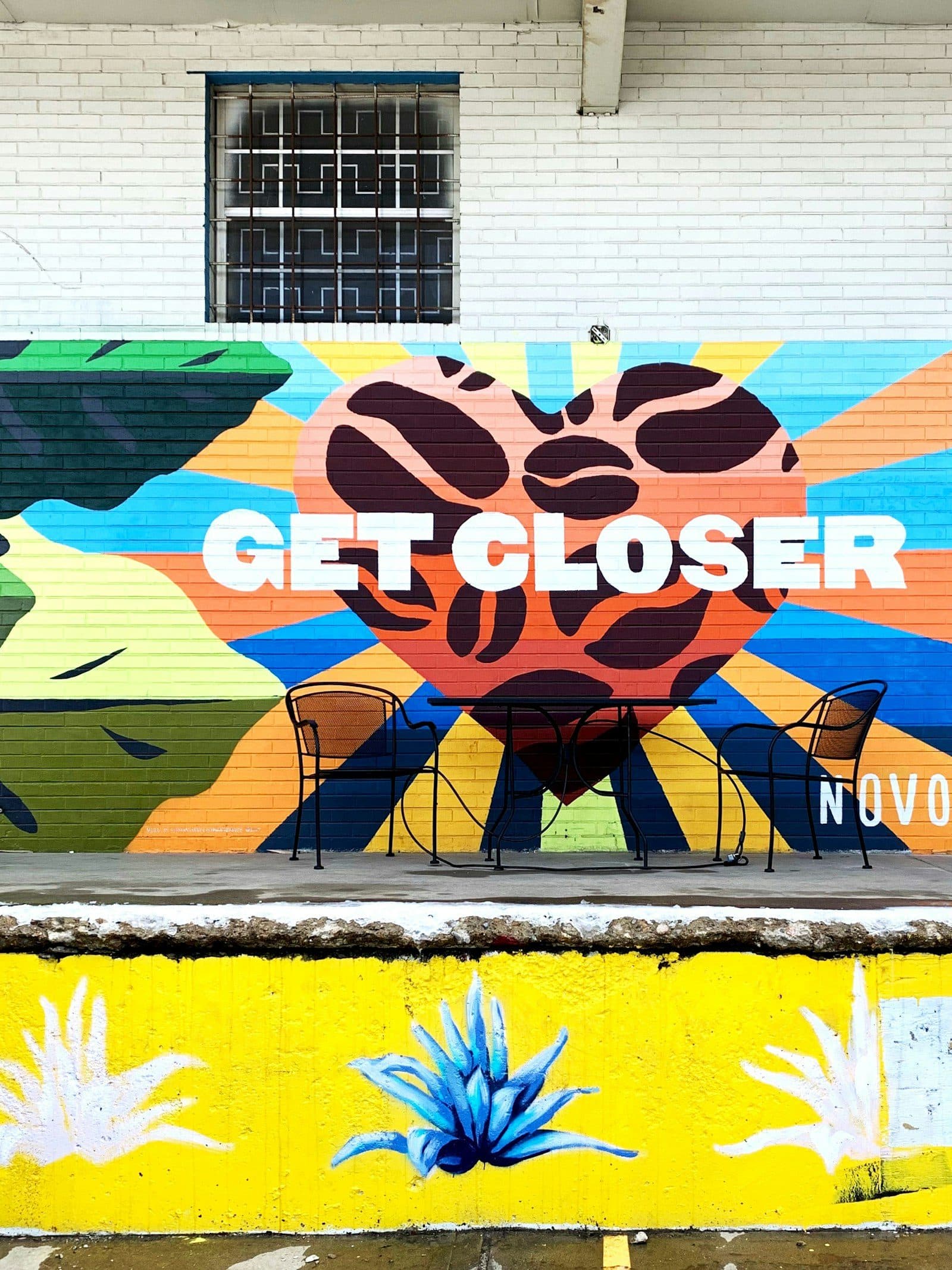 A bright 'Get Closer' mural beside a small cafe table outside a RiNo coffee shop