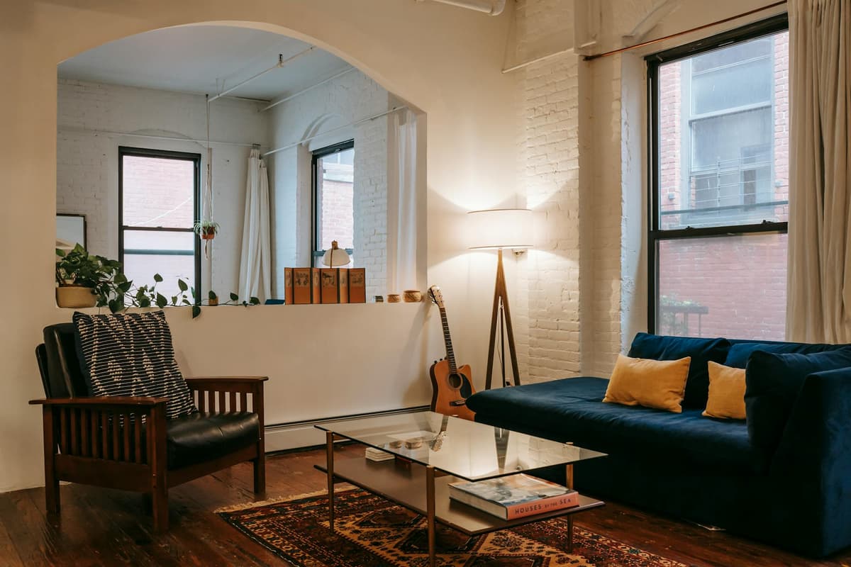 A warm, furnished apartment living room with a navy sofa, exposed brick, and natural light