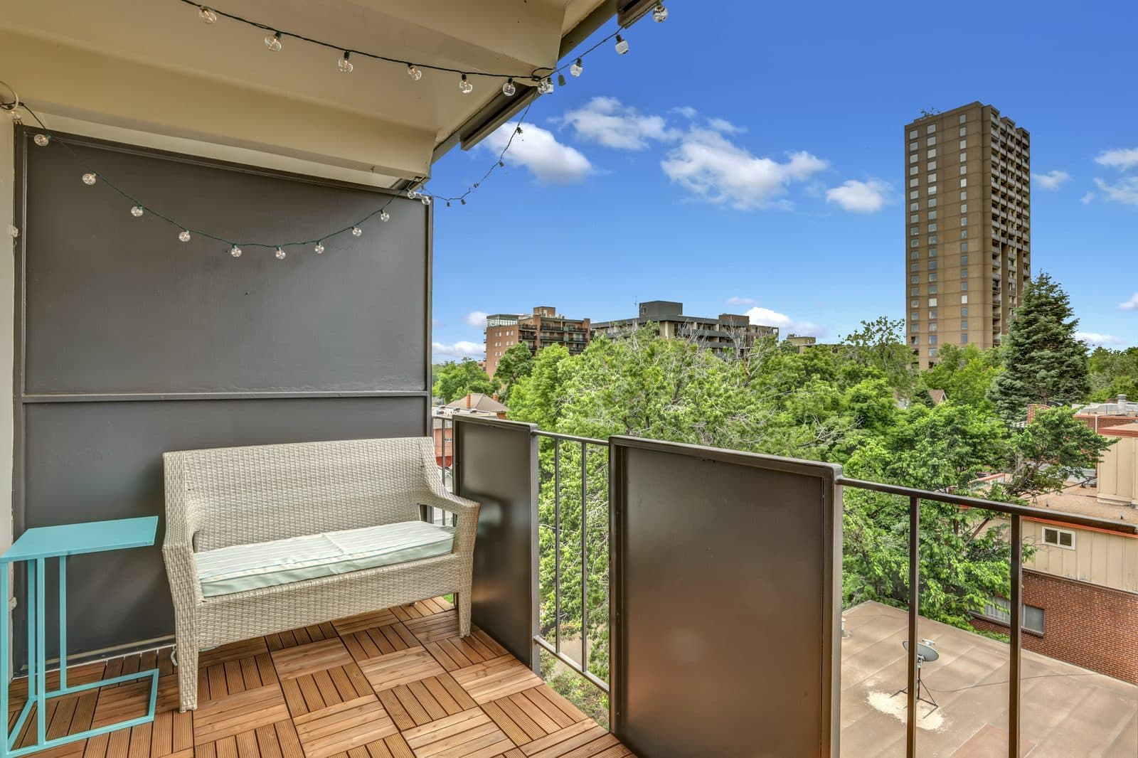 Balcony with wicker bench, string lights and a view of the residential tower across the park